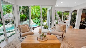 A spacious living area with large windows, a wooden coffee table, two beige armchairs, and a hammock. - Heated pool / Lush Tropical Surrounding / Castaway Key / RESlDENCES (Fort Lauderdale)