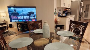 Un comedor con una mesa de vidrio rodeada de sillas de madera con cojines de asiento trenzados, un televisor mostrando la interfaz de un servicio de streaming, y una cocina con armarios y un refrigerador al fondo.