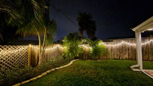 Un patio trasero con cerco de madera, luces colgantes y un pequeño jardín con palmeras y arbustos.