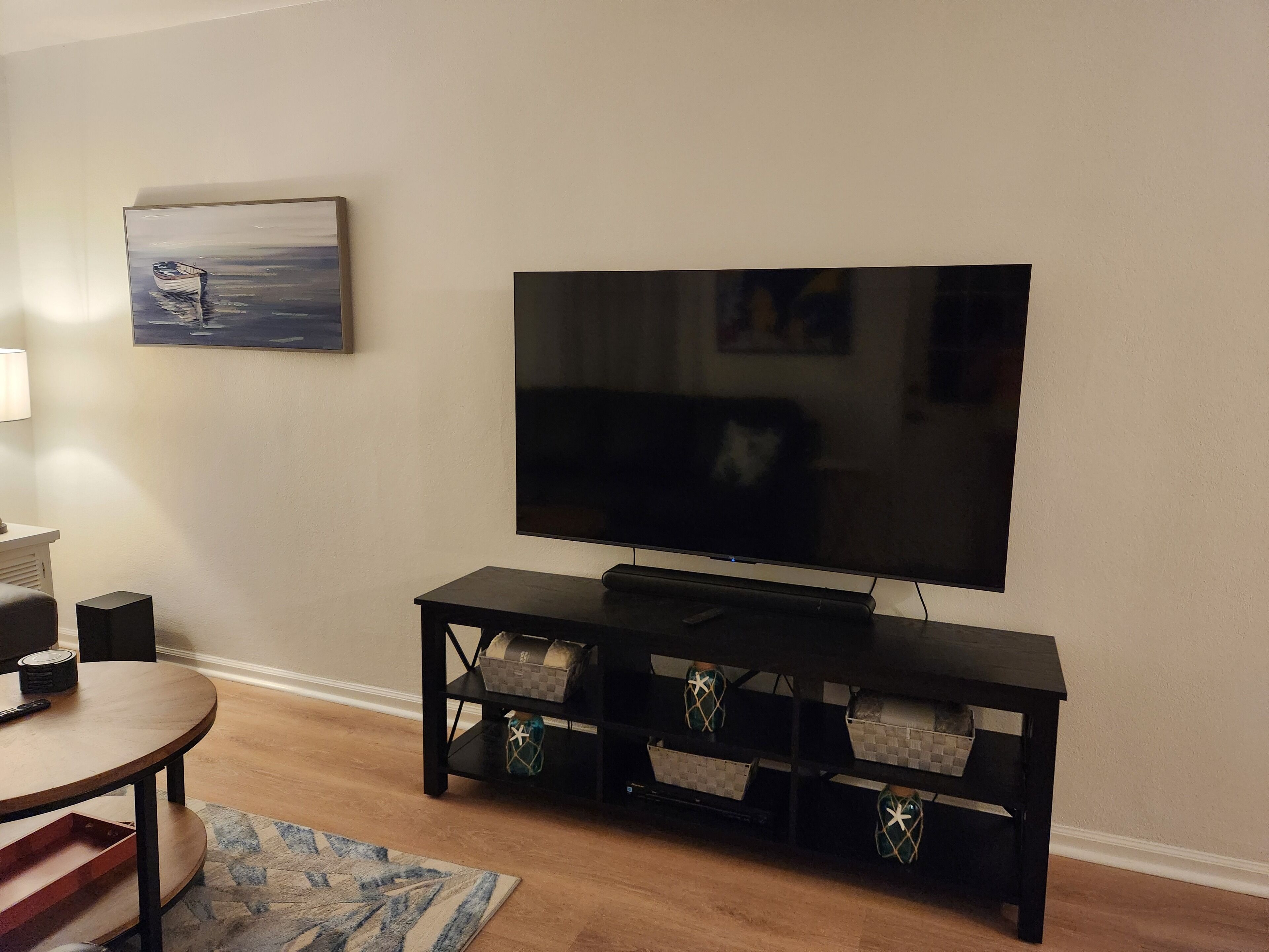 A living room with a flat-screen TV on a black stand, a painting on the wall, a small table with a lamp, and a patterned rug.