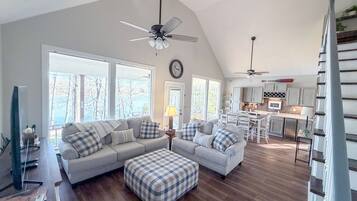 A spacious living area with a large sectional sofa, a matching ottoman, and a dining table with chairs. There are two ceiling fans, a kitchen with white cabinets, and large windows offering a view of trees.