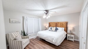 Une chambre à coucher comprenant un lit, une chaise, une table de chevet, un ventilateur de plafond et une fenêtre avec des rideaux.
