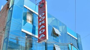 A modern hotel with a blue glass facade, a prominent red sign, and multiple flags. - Hostal Mansion Dorada (Tacna)