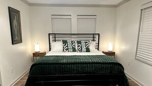 A bedroom with a bed, two bedside tables, a nightstand, a framed picture on the wall, and a window with blinds.