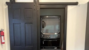 A kitchen with a black door, stainless steel appliances, and a small trash bin.