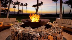A fire pit with a stone base surrounded by lounge chairs, set against a backdrop of palm trees and a sunset. - Ocean Front Westin Villas North!  Gorgeous Pools, Beach & Amenities! (Lahaina)