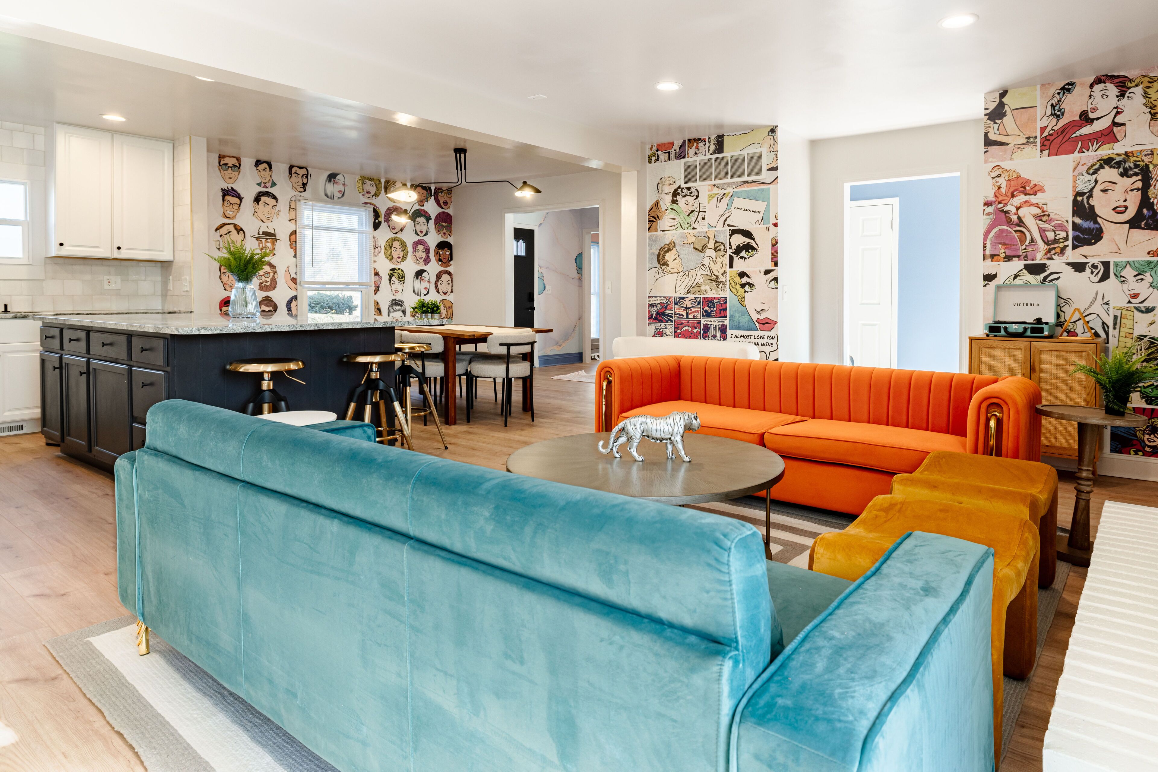 Una sala de estar moderna con un sofá color turquesa, un sofá naranja y un reposapiés amarillo. Hay una zona de cocina con armarios blancos y una mesa de comedor. Las paredes están decoradas con ilustraciones al estilo de cómic.