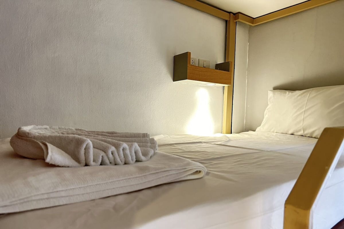 Una cama bien hecha con una toalla doblada, un cabecero de madera y una mesita de noche con una lámpara.