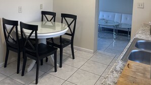 Een eetgedeelte met een ronde tafel en vier stoelen, een keuken met een spoelbak en aanrecht, en een blauwe wand met een bankje.