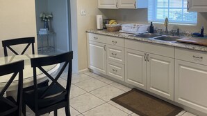 Een keuken met witte kastjes, een granieten aanrechtblad, een raam met zonwering, en een eethoek met tafel en stoelen.