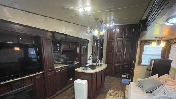 Cocina compacta con estufa integrada, refrigerador pequeño y lavamanos. En la zona de estar, hay un otomán de cuero y un sofá.