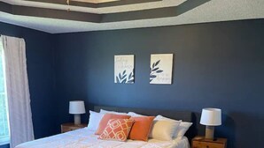 Une chambre à coucher comprenant un lit, deux tables de chevet avec des lampes, un ventilateur de plafond et deux œuvres d’art murales.