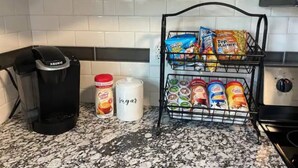 Une cuisine équipée d’une machine à café Keurig, d’un récipient de crème à café, d’un sucrier et d’un présentoir contenant divers aliments, notamment des en-cas et des dosettes de café.
