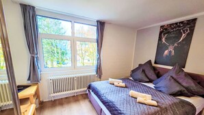 Ein Schlafzimmer mit großem Fenster, einem Bett mit aufgerollten Handtüchern, einem Holz-Nachttisch und einem Hirschgemälde an der Wand.