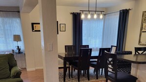 Un espace repas doté d’un parquet en bois disposé en motif chevron, d’une table à manger avec des chaises, d’un lustre et d’une vue sur l’extérieur grâce à de grandes fenêtres équipées de stores.