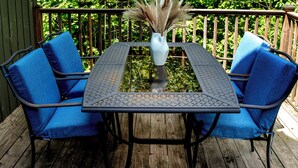 A wooden deck with a metal table and two blue chairs, surrounded by lush greenery.