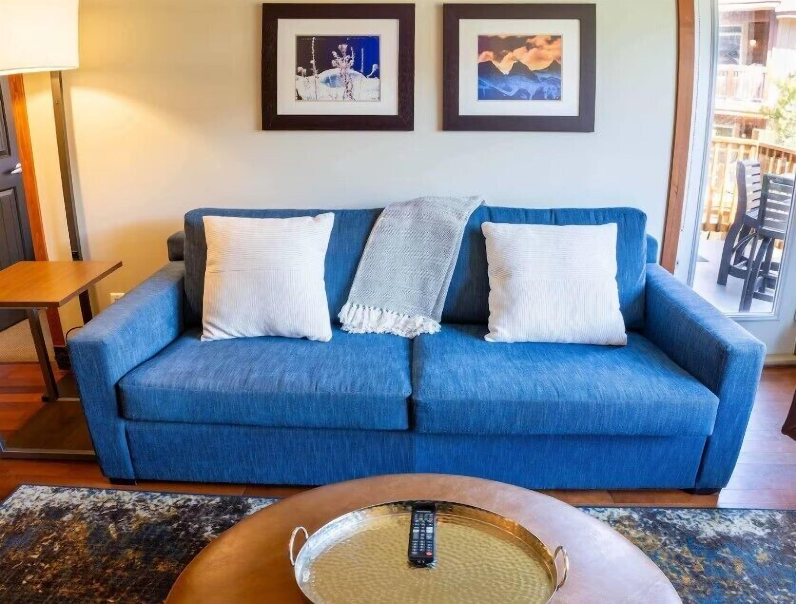 A blue sofa with white pillows and a throw blanket, a wooden coffee table with a remote control, and two framed pictures on the wall.