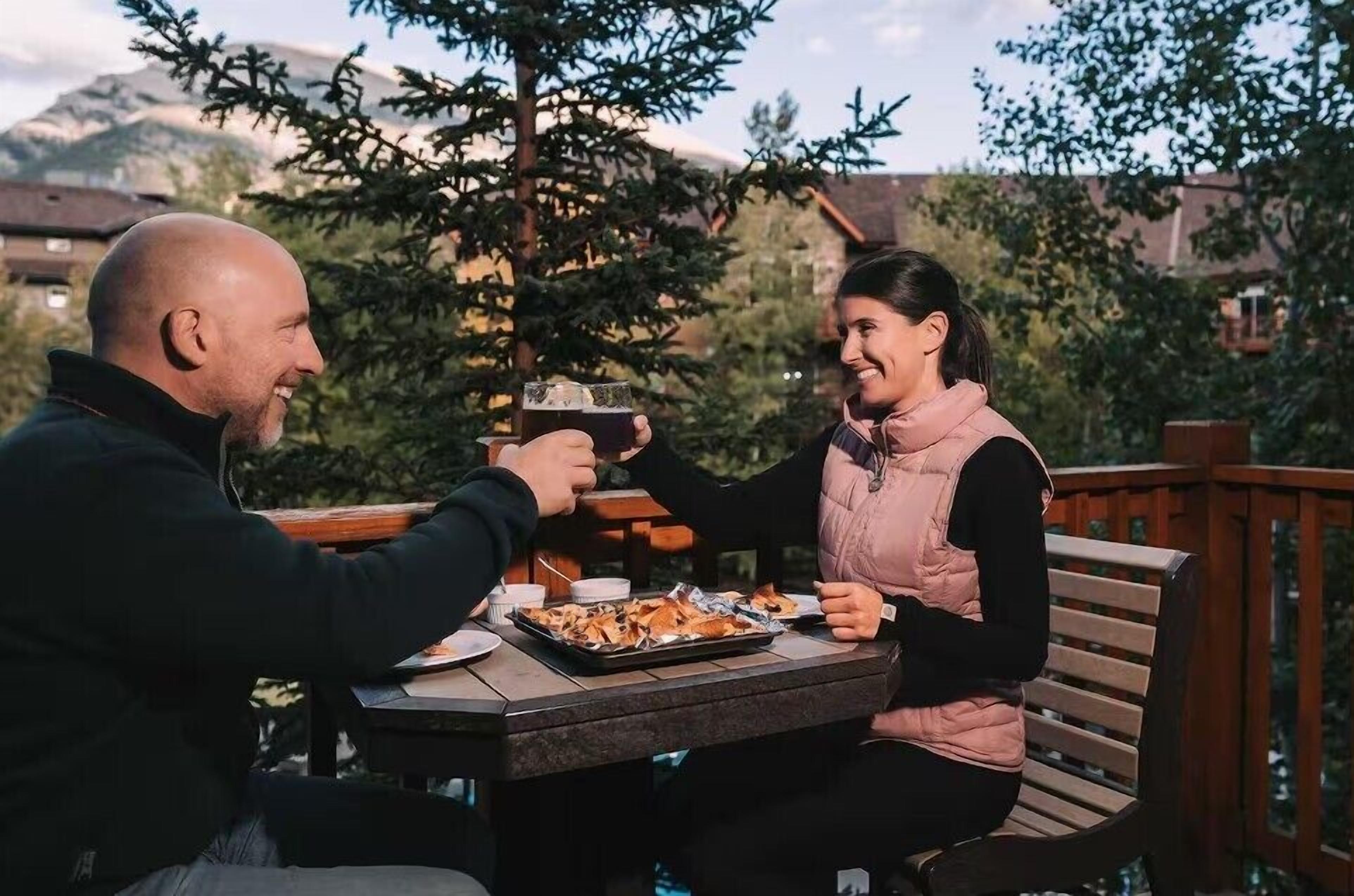 Duas pessoas sentadas numa mesa ao ar livre, com uma bandeja de comida e bebidas, com vista para uma paisagem montanhosa.