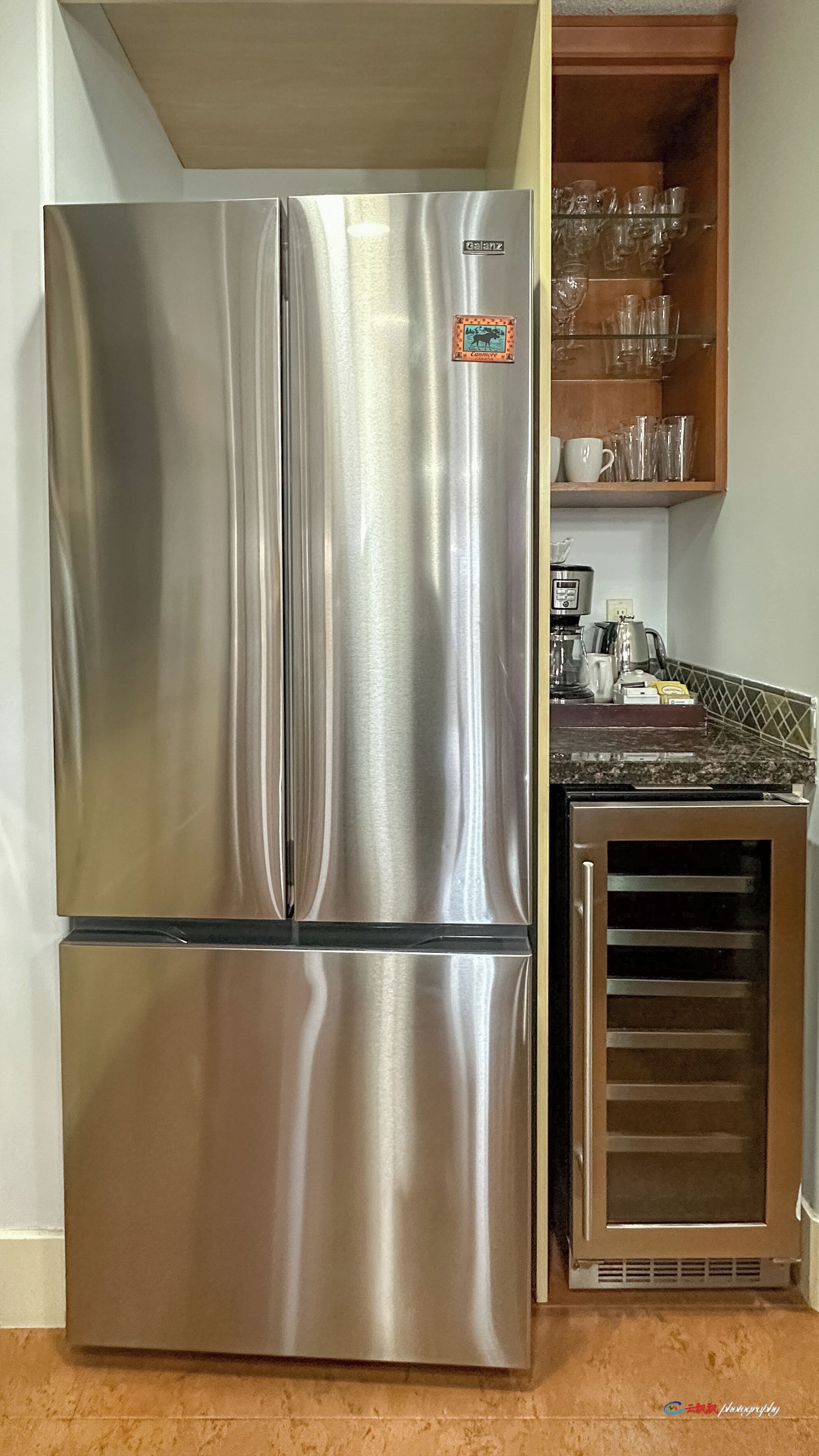 A stainless steel refrigerator with a digital display, a wooden cabinet with glassware, and a granite countertop.