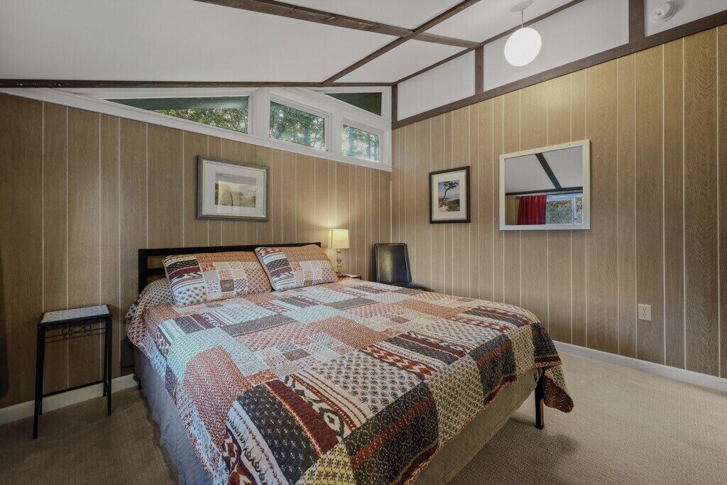 Ein Schlafzimmer mit einem großen Bett, einem Nachttisch, einer Wandlampe und einem Fenster mit Blick auf Bäume.