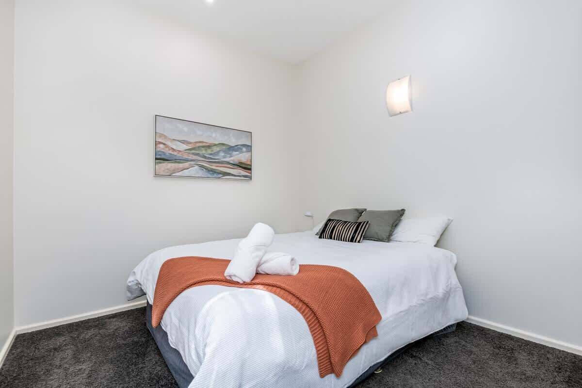 A neatly made bed with white linens and a brown throw blanket, a painting on the wall, and a single bedside lamp.