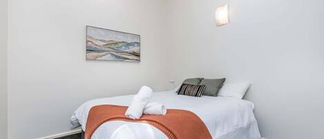 A neatly made bed with white linens and a brown throw blanket, a painting on the wall, and a single bedside lamp.