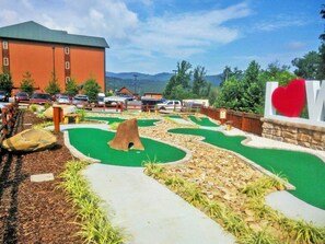 A miniature golf course with a green putting area, a rock path, and a large building in the background.