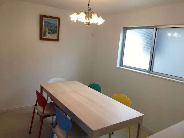 Un coin repas avec une table en bois et des chaises colorées, un lustre, un tableau au mur et une fenêtre avec des stores.