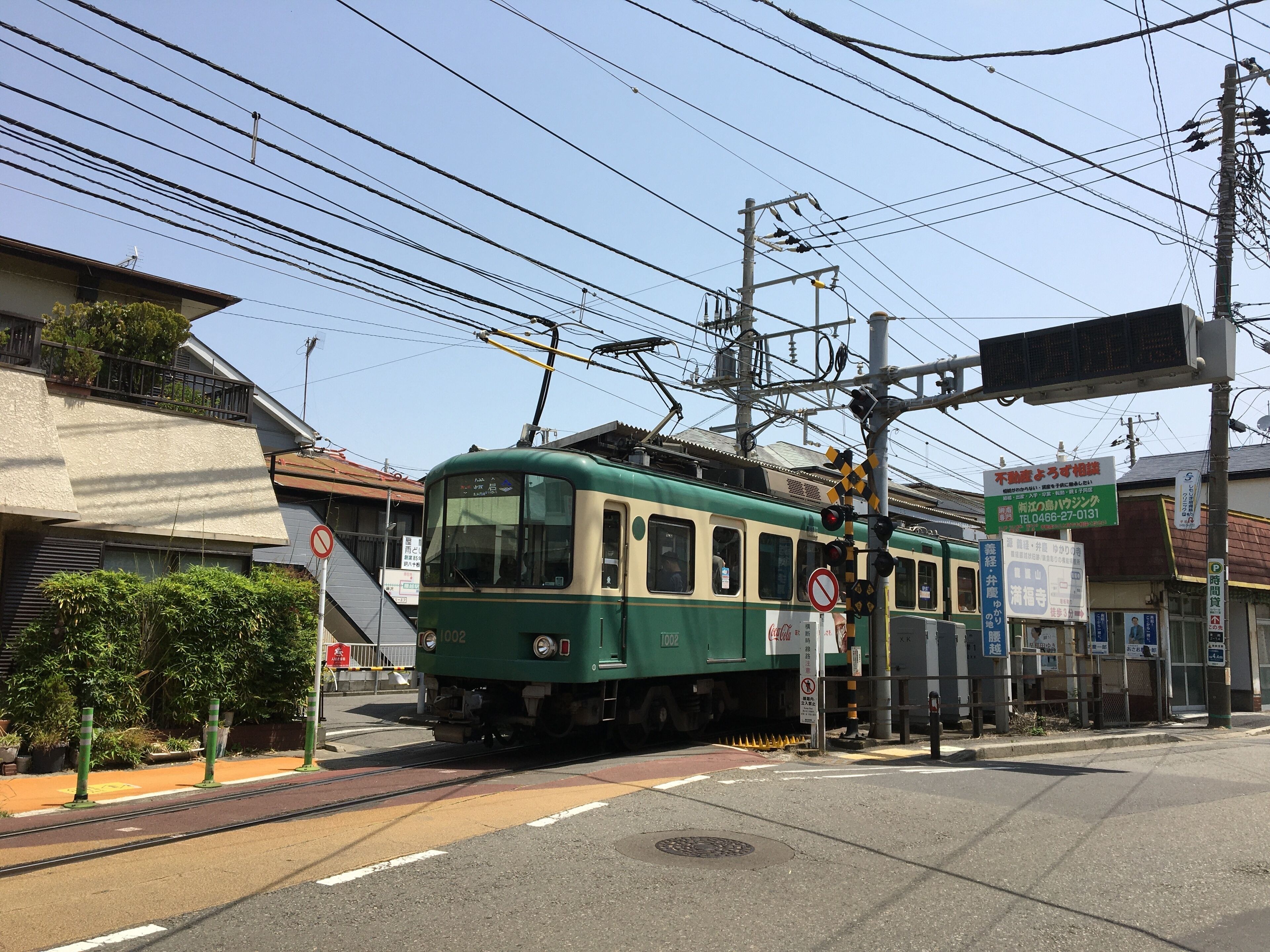 A green tram at a station with overhead wires and a pedestrian crossing.
