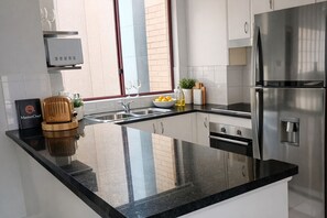 A modern kitchen with a black countertop, stainless steel appliances, a built-in oven, and a window with blinds.