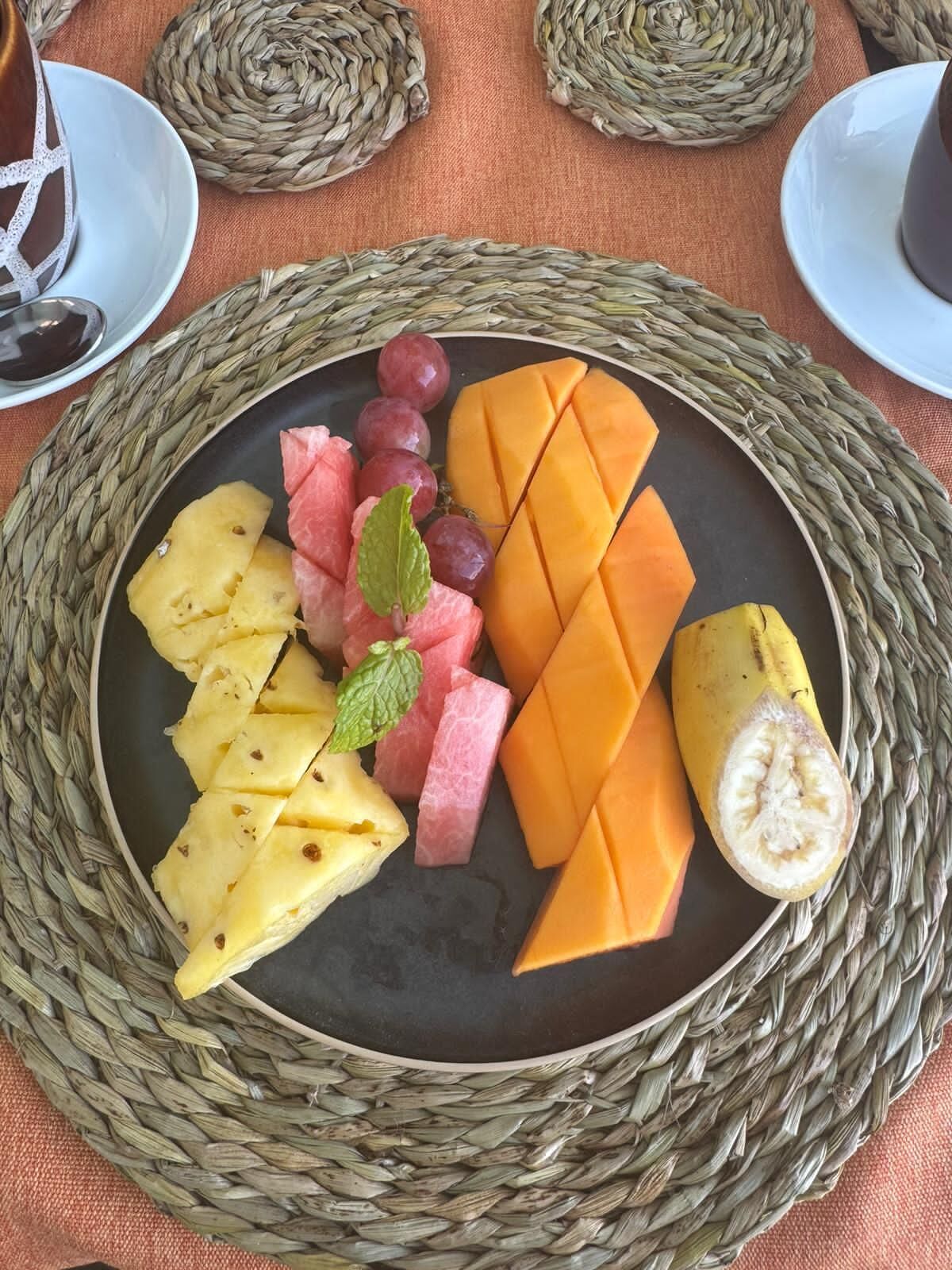 A plate of fruit including pineapple, watermelon, grapes, and mango slices, surrounded by woven placemats.