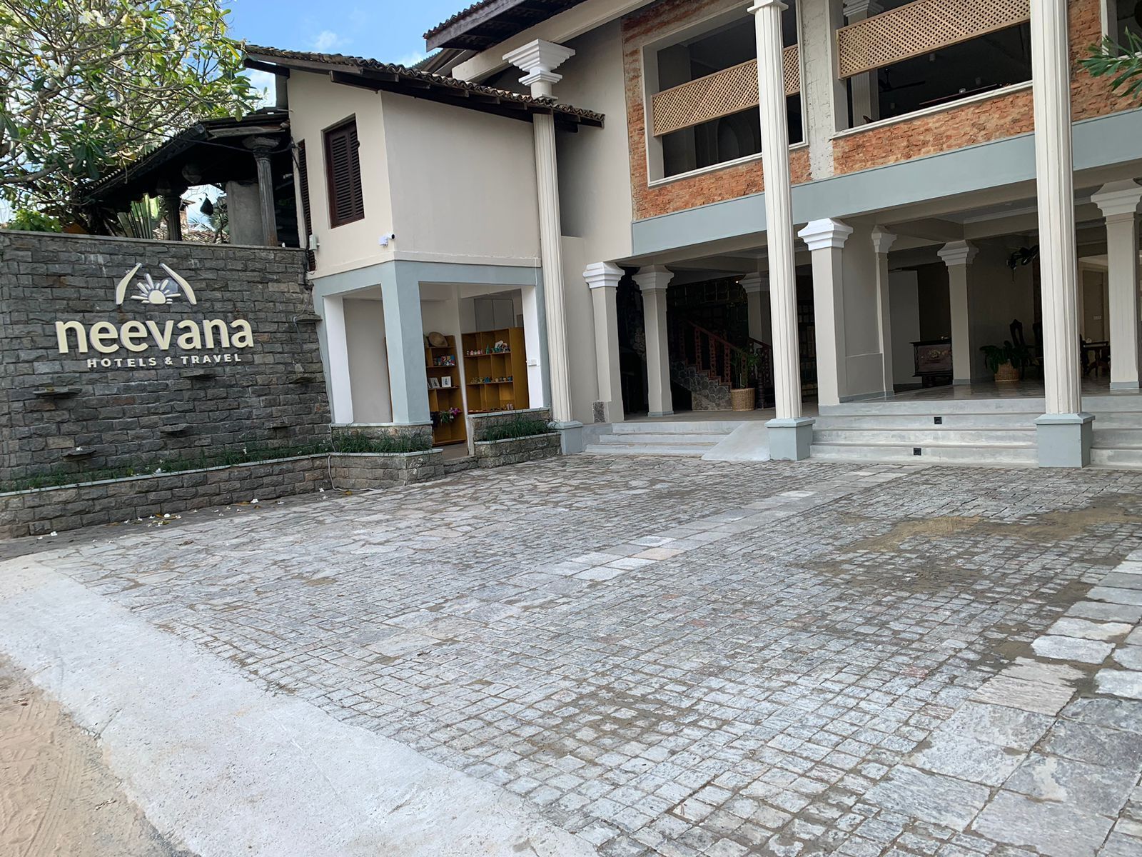A hotel entrance with a stone-paved driveway, a sign for Neevana Hotels & Travel, and a covered entrance area with columns.