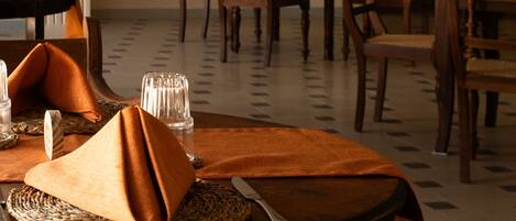 A dining area with tables set for two, featuring woven placemats and orange napkins.