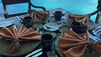Una mesa de comedor dispuesta con platos, cubiertos y individuales tejidos, con vistas a una piscina y al océano.