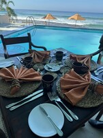 Una mesa de comedor dispuesta con platos, cubiertos y individuales tejidos, con vistas a una piscina y al océano.