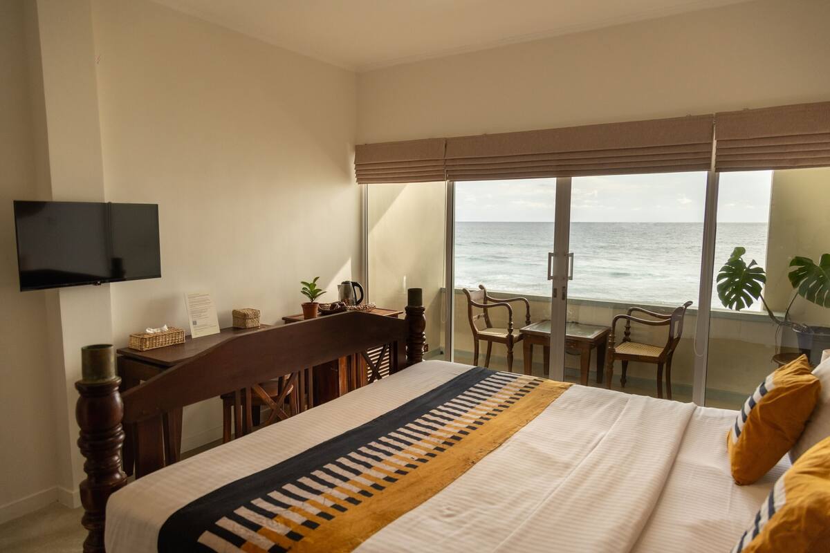 Ein Hotelzimmer mit einem Bett, einem an der Wand befestigten Fernseher, einem Balkon mit Meerblick und einem Tisch mit Stühlen.