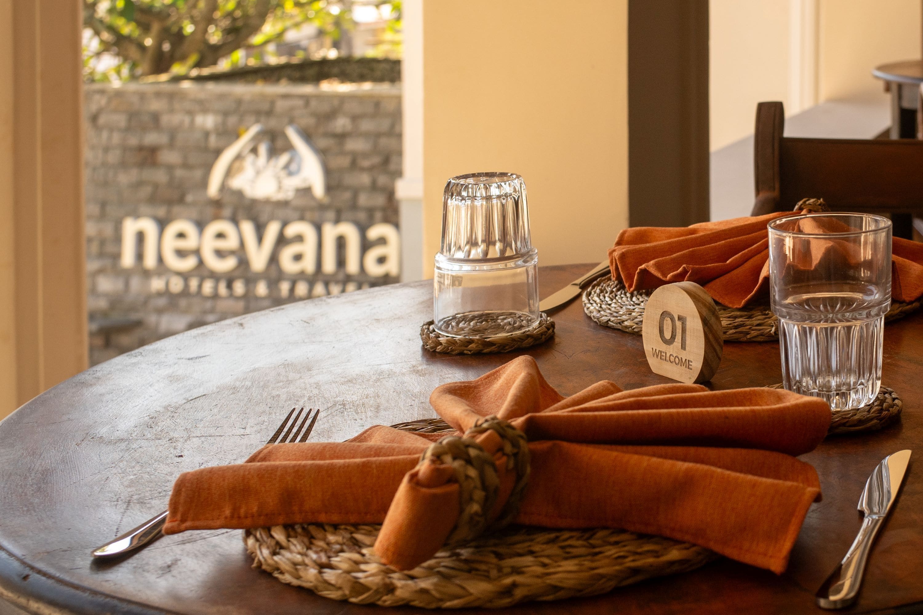 A table set with a woven placemat, a folded orange napkin, a glass of water, a glass of water, a salt shaker, and a fork and knife on a wooden table.