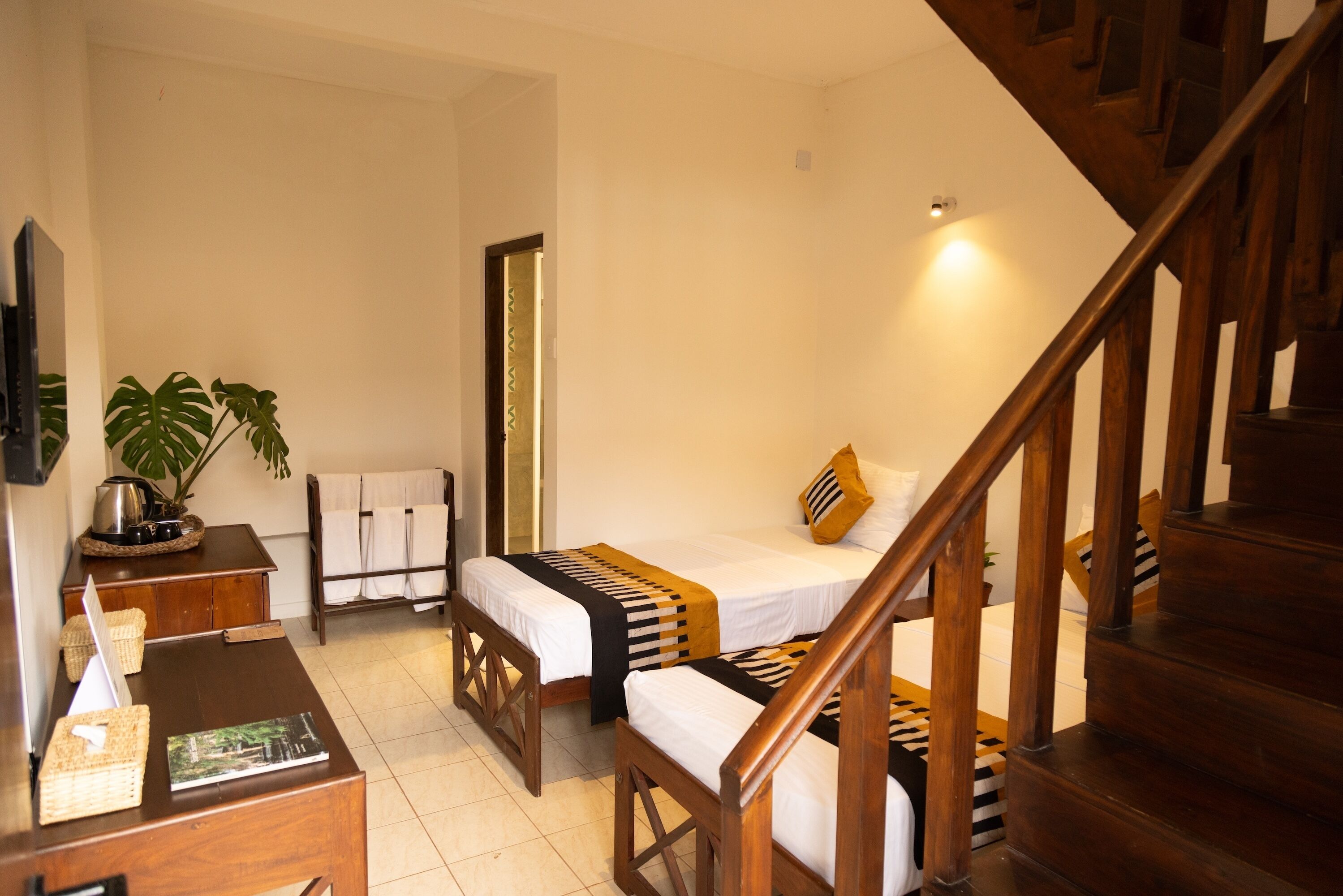 A hotel room with a wooden staircase, two beds with striped bedding, a wooden desk, a plant, and a television.
