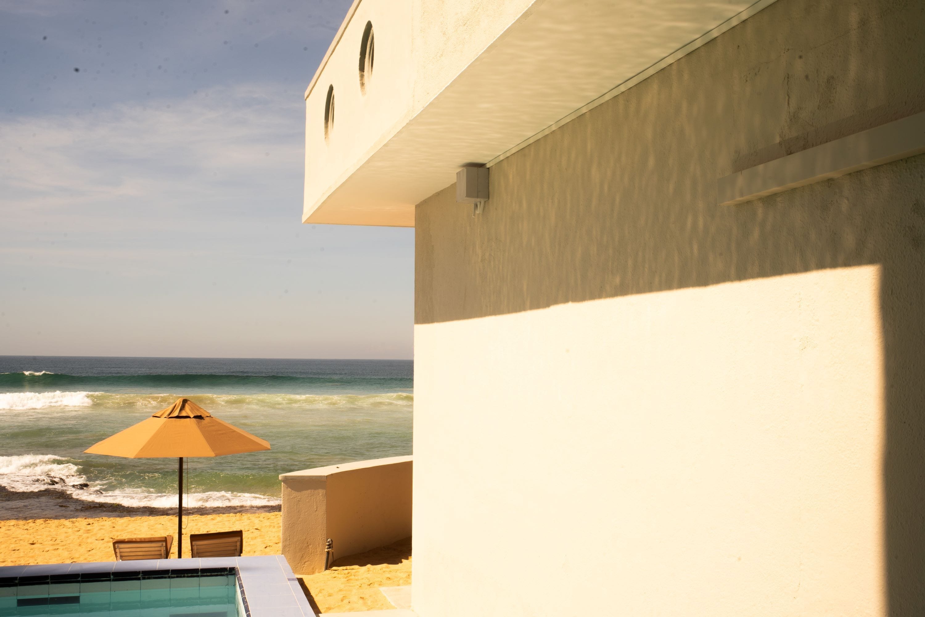A pool area with a view of the ocean, a beach umbrella, and a building with a light-colored exterior.