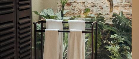 A room with a bed, a window with a view of a brick wall and plants, and a towel rack with towels.