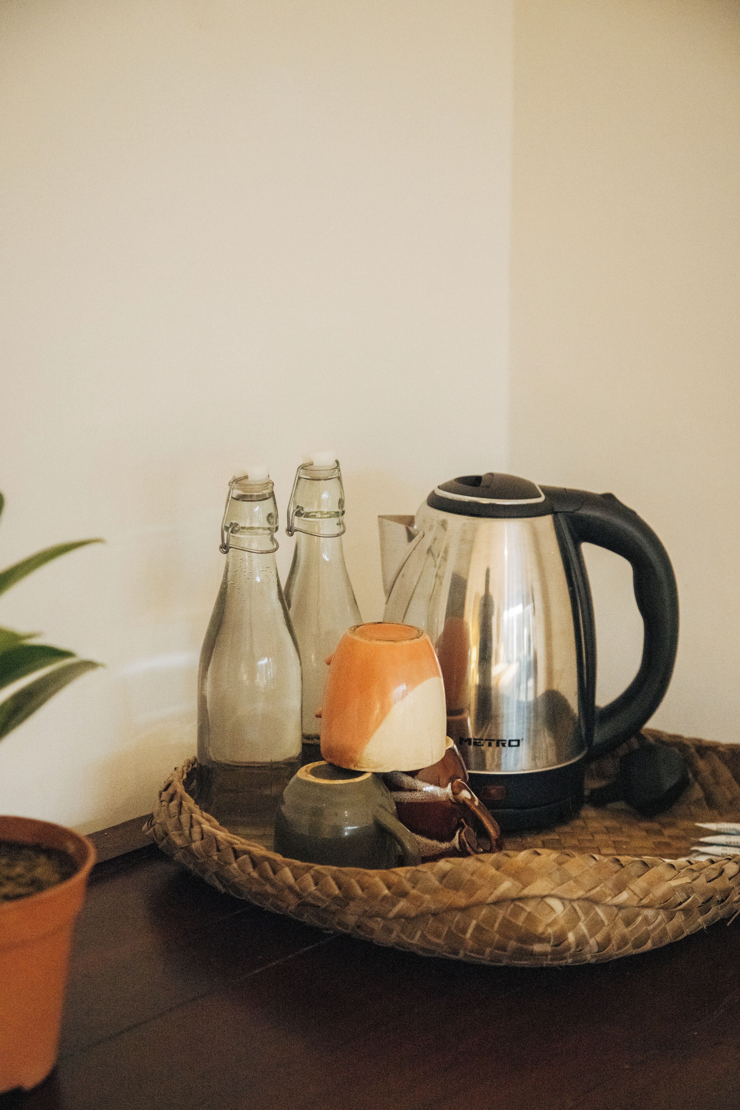 A kitchen counter with a kettle, two bottles, a mug, and a woven tray.