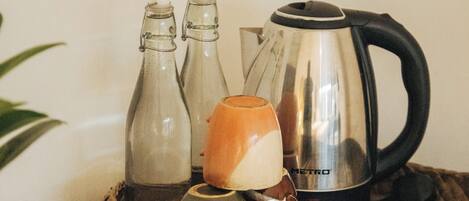 A kitchen counter with a kettle, two bottles, a mug, and a woven tray.