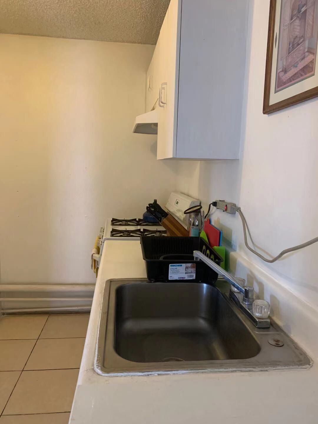 Una piccola cucina con un lavello in acciaio inossidabile, un piano di lavoro bianco e un pensile sopra il lavello. Sul piano di lavoro si trovano diversi utensili da cucina e un espositore per piatti.