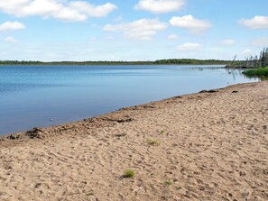 En sandstrand med en lugn sjö och en skog i bakgrunden.