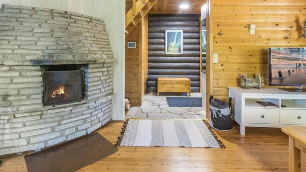 A log cabin interior with a stone fireplace, wooden floors, a staircase, and a television on a white cabinet.
