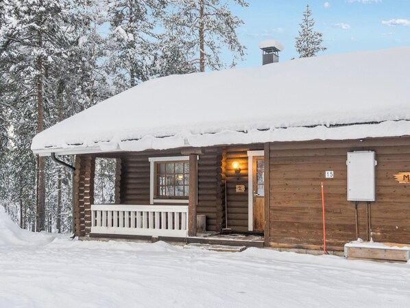 Lumipeitteinen puinen mökki, jossa on kuisti ja savupiippu.
