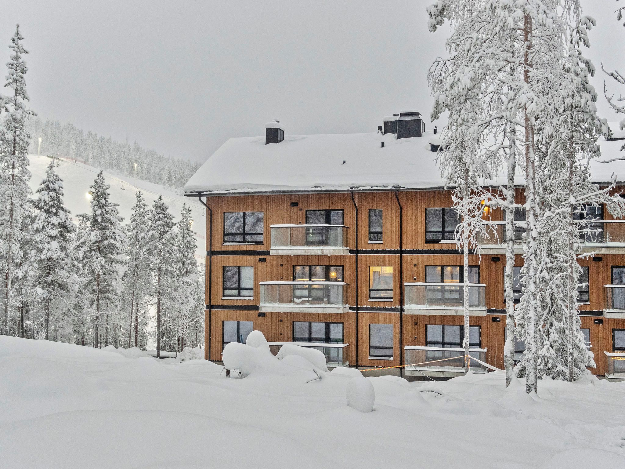 Un immeuble en bois à plusieurs étages, doté de balcons, entouré d’arbres couverts de neige et avec une piste de ski en arrière-plan.