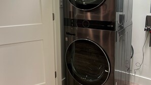 A compact, stainless steel front-loading washer and dryer unit in a corner of a room with wooden flooring and white walls. - Cozy Cascade Nook - 2 bedroom basement apartment (OREM)
