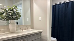 A bathroom with a white sink, a mirror, a vase with green plants, and a dark blue shower curtain. - Cozy Cascade Nook - 2 bedroom basement apartment (OREM)