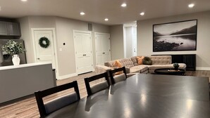 A modern living room with a large dining table, a sofa, and a kitchen area with cabinets and a refrigerator. - Cozy Cascade Nook - 2 bedroom basement apartment (OREM)